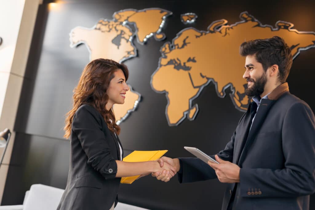 Two business professionals shake hands and smile at each other in an office with a large world map on the wall behind them; one holds a yellow folder, the other a tablet.