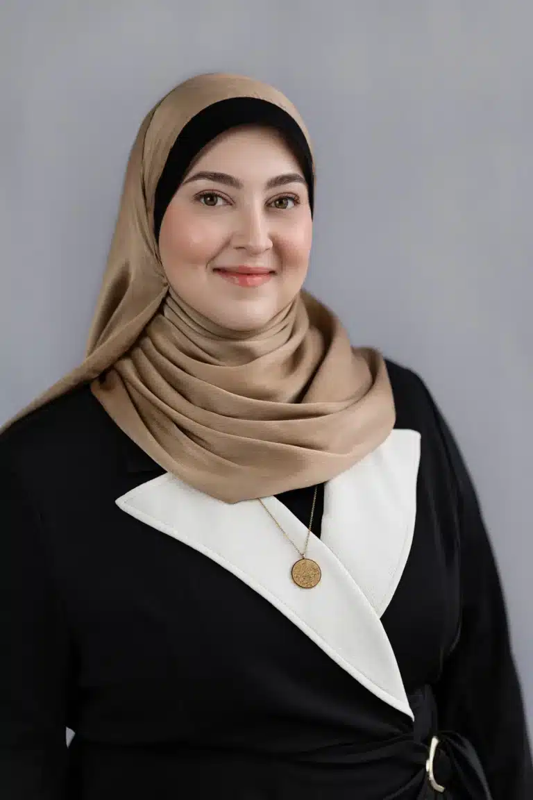 Mariam Jammal, wearing a beige hijab and a black outfit with a white accent, smiles at the camera against a plain gray background. She has on a gold necklace and showcases a natural makeup look.