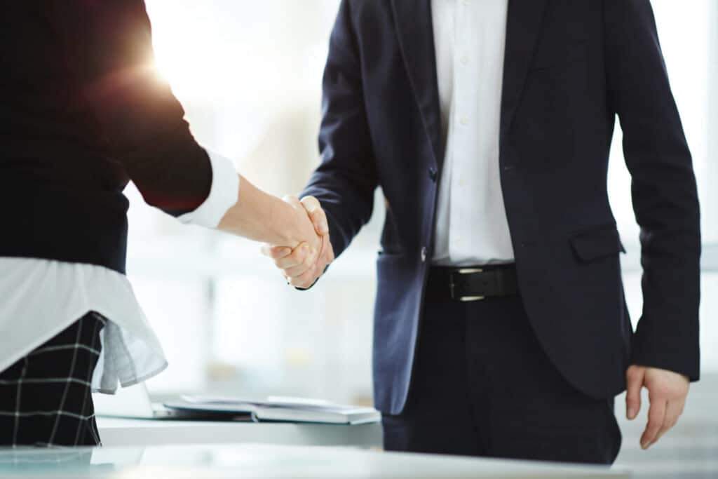 Two people in business attire shaking hands in an office setting, symbolizing agreement or partnership. Only their torsos and hands are visible. Sunlight is shining in the background.