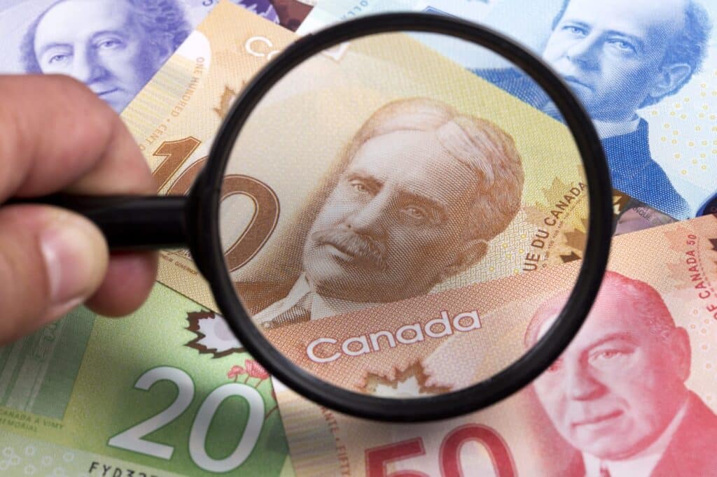 A hand holds a magnifying glass over Canadian dollar bills of various denominations, focusing on the portrait on the ten-dollar note. Other colorful banknotes, including $20 and $50, are visible underneath.