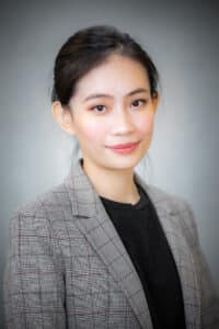A young woman with long dark hair tied back, wearing a gray plaid blazer over a black top, smiles softly against a plain, blurred gray background.