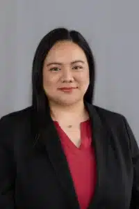 Niña Catacutan, with long black hair, wears a black blazer over a red blouse and a gold necklace, standing in front of a plain gray background and smiling slightly at the camera.