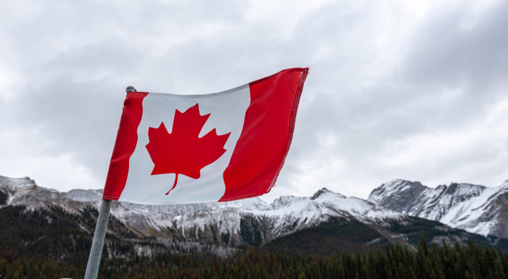 The Canadian flag flying proudly against snow-capped mountains and cloudy skies, representing the strong business environment and economic opportunities that attract entrepreneurs and investors seeking to establish operations in Canada's stable market, where companies can access financing, evaluate investment opportunities, and benefit from the country's robust financial system and business-friendly policies that support both domestic and international commerce across diverse industries and sectors.