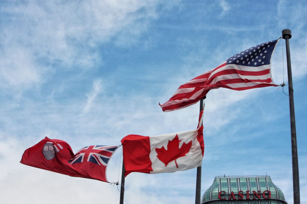 Here's a natural alt text for this image using terms from the new list: "American and Canadian flags flying together against a blue sky, symbolizing the cross-border relationship for US citizens seeking to move to Canada through various pathways including the Express Entry System, Federal Skilled Worker Program, and Provincial Nominee Programs, where skilled workers can obtain work permits, study permits, or pursue permanent resident status through the Canadian immigration system while navigating considerations like Canadian taxes, housing costs, universal healthcare, and provincial health insurance as they transition from temporary residents to permanent residents under the Canadian government's programs administered by Citizenship Canada.