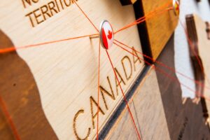 A close-up of a wooden map featuring the word CANADA engraved, with red strings attached to a pin marked by a small Canadian flag—an inspiring symbol of business opportunities in Canada for foreigners. The surrounding area includes more labeled wood pieces.