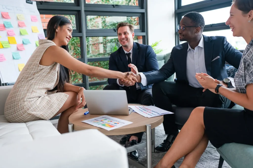 Four business professionals sit around a table, two shaking hands and smiling—perhaps celebrating a deal to buy a small business in Canada. Laptops, papers, and charts cover the table beneath a whiteboard filled with sticky notes.