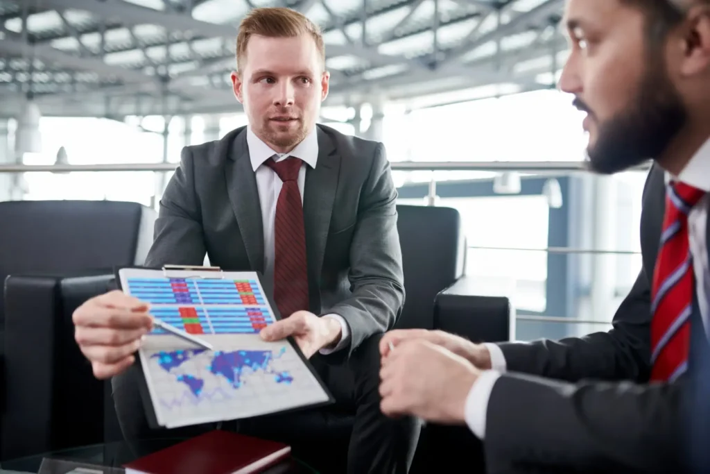 Two businessmen in suits discuss data; one holds a tablet showing colorful charts and maps, possibly analyzing business immigration trends or buying a business opportunities in a modern office setting.