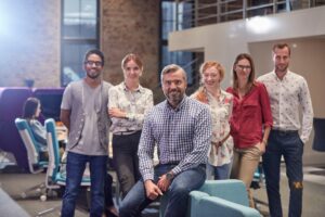 Six people pose and smile in a bright, modern office space, reflecting a diverse team that could thrive through immigration for technology in today's global workforce. Both men and women are casually dressed, with desks and coworkers in the background.