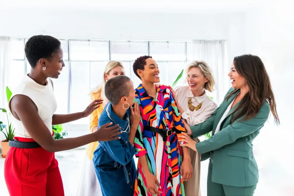 Five women stand together indoors, smiling and laughing. Dressed in colorful, stylish outfits, they enjoy each other’s company in a bright, modern room—celebrating successes and connections through business immigration opportunities.