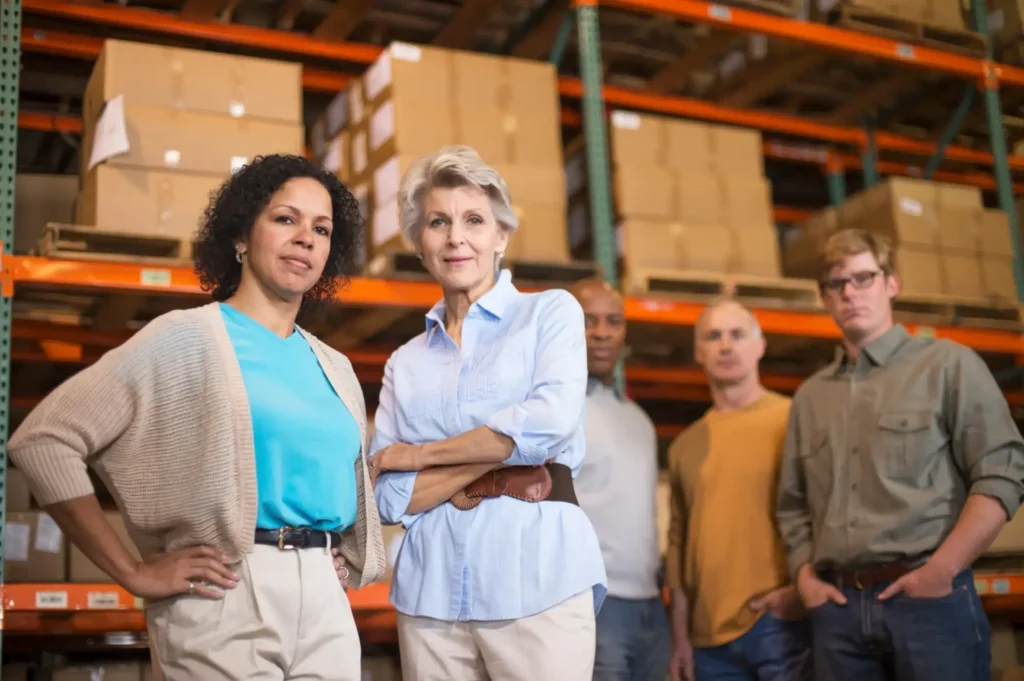 Five adults stand confidently in a warehouse with shelves of boxes behind them. This diverse team, dressed in business casual, exemplifies the workforce supported by Labour Market Impact Assessment (LMIA) and Work Permit processes for foreign investors.
