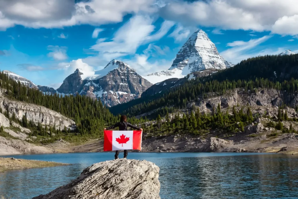 A person stands on a rock by a lake, holding a Canadian flag, with pine trees and snow-capped mountains under a blue sky—capturing the dream of newcomers seeking opportunities through LMIA high wage programs in Canada.