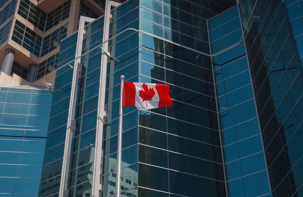 A Canadian flag waves on a flagpole in front of a modern glass building, its blue-tinted windows reflecting the cityscape—an inviting scene for foreign investors exploring opportunities in Canada.