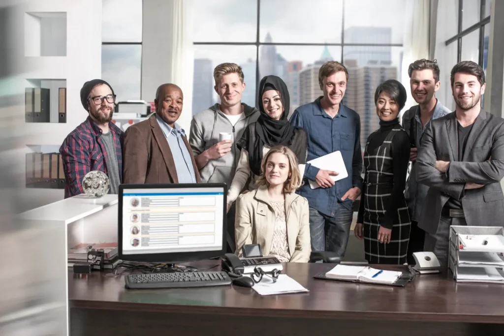 A diverse group of nine people, men and women of various ages and backgrounds, stand and sit smiling in an office setting behind a desk with business visa paperwork. Large windows reveal a city skyline.