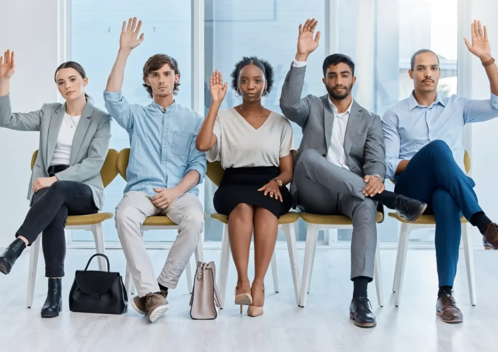 Five professionally dressed people sit in a row on chairs, each raising a hand as if responding to a question about high wage LMIA requirements, with neutral expressions in a bright, modern office setting.