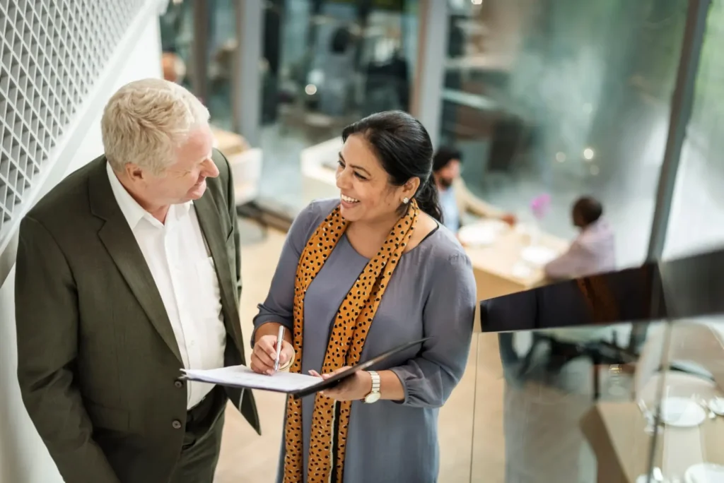 A man in a suit and a woman in business attire with a patterned scarf are smiling and discussing High wage LMIA requirements on a clipboard. They appear to be in a modern office with people in the background.