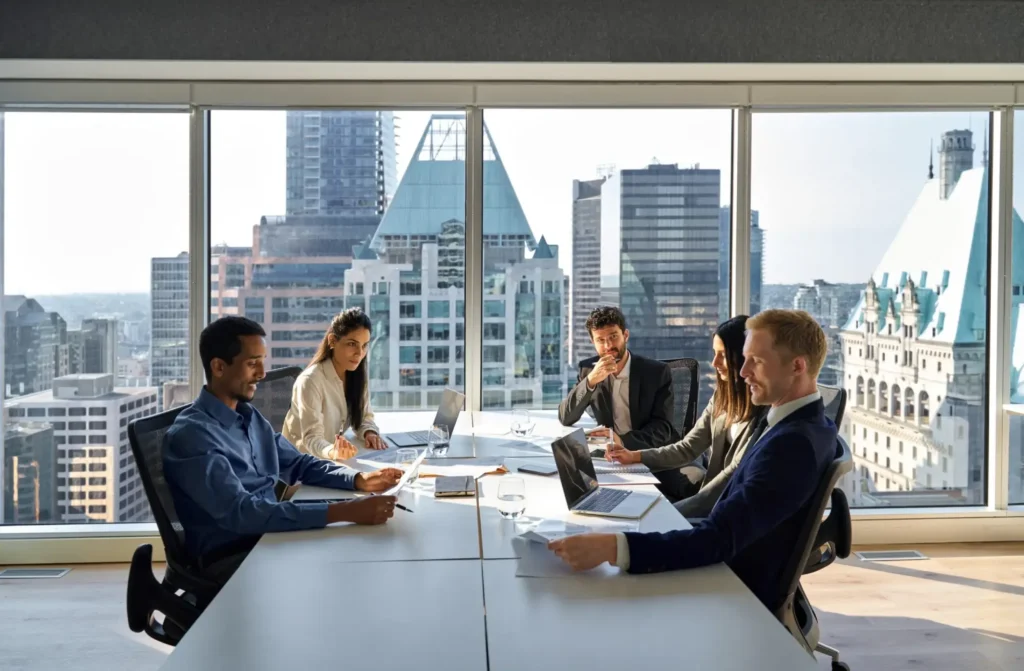 Six people sit around a conference table in a modern office with large windows and city views, engaged in a business meeting discussing business immigration opportunities for foreign investors buying a business, using laptops and documents.