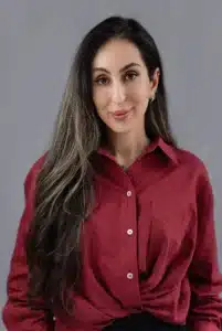 A woman with long, wavy brown hair wearing a red button-up shirt stands in front of a plain gray background, smiling softly at the camera.