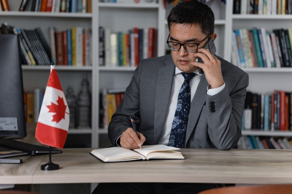 Canadian business immigration lawyer Rakhmad Sobirov speaks on his cell phone while taking notes at his desk, offering expert legal guidance on the Express Entry system, skilled worker streams, the Ontario Immigrant Nominee Program, the Nova Scotia Nominee Program, the Alberta Advantage Immigration Program, and BC PNP, helping international graduates, entrepreneurs, and skilled workers navigate eligibility requirements, prove language ability, obtain educational credential assessments, and succeed in Canada’s competitive federal immigration programs.
