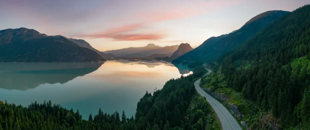 A stunning highway winding along a pristine lake and towering mountains in British Columbia, Canada, symbolizing the natural beauty and economic potential driving the BC Provincial Nominee Program (BC PNP), which draws skilled workers, international students, and construction sector professionals looking to navigate the Express Entry pool, prove language ability, and meet provincial labour market demands in a thriving Canadian province.