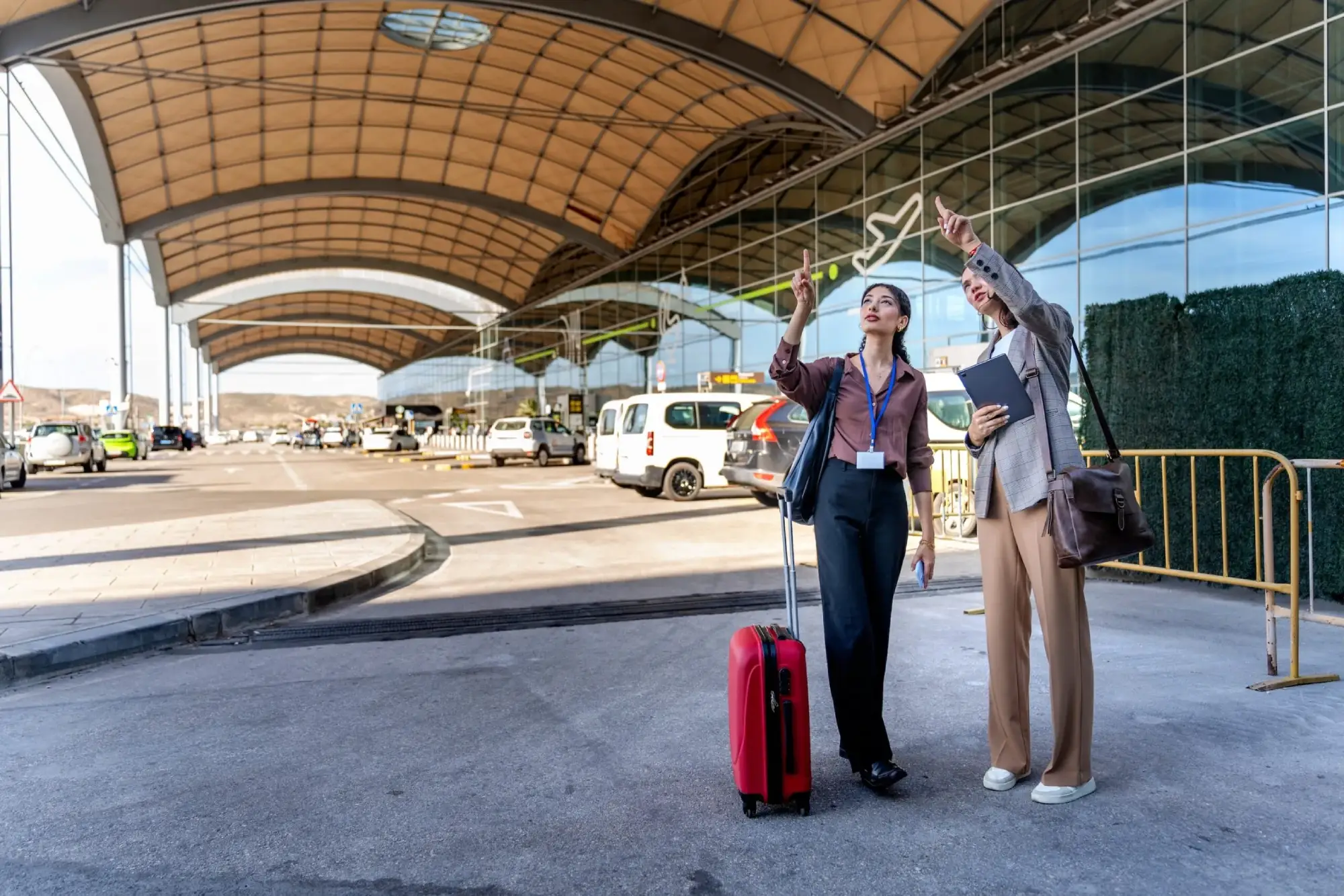 Two businesswomen with luggage pointing towards the sky outside a modern airport terminal.