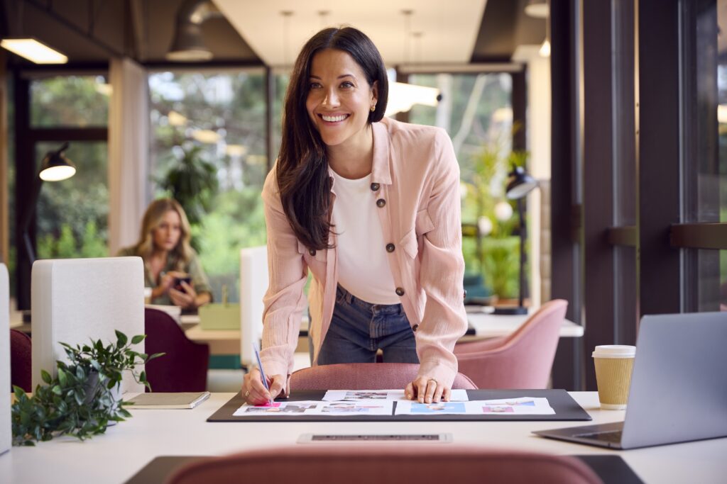 Smiling business owner in a pink blazer standing at her desk in a modern office, reviewing detailed business plans and supporting documents as part of the application process for the Provincial Nominee Program, demonstrating her solid business plan and proven track record while working with a regulated Canadian immigration consultant to navigate business immigration programs, achieve business milestones, and establish business operations that will provide significant economic benefits through creating jobs for local tech professionals and supporting local initiatives in the Canadian economy.