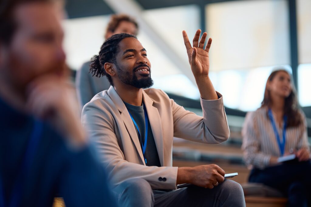 Enthusiastic entrepreneur raising his hand during a business conference, representing self employed professionals who bring market research expertise and seek professional guidance for their Start Up Visa application, demonstrating success in proving genuine intent for business viability through their gourmet food business model that requires a labor market impact assessment, business revenue statements, and financial records while partnering with Canadian suppliers and contributing social or cultural contributions to achieve business ownership and support seasonal businesses through their consultancy or freelance operation under Citizenship Canada programs.