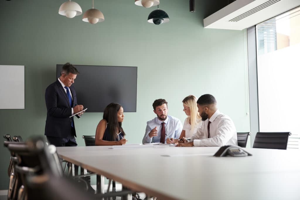Five people in business attire are in a meeting room discussing an ICT work permit. Four are seated at the table, engaged in conversation, while one stands taking notes. A large screen and hanging lights are visible in the background.