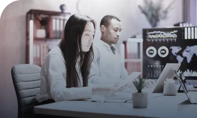 Two people sit at a desk in an office, focused on their work. The woman reviews investment immigration data on her computer, while the man works on his laptop. Charts and a world map are displayed on a monitor in the background.