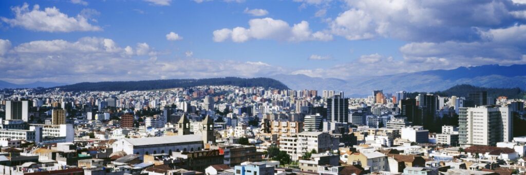 Panoramic view of a city with numerous modern buildings under a blue sky, ideal for professionals on an Intra Company Transfer, with mountains in the background and sunlight illuminating the urban landscape.