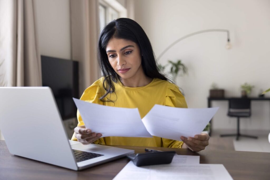 A woman in a yellow top sits at her desk, focused on reviewing two sheets of paper—possibly related to her C11 Work Permit. Her laptop, calculator, and additional papers are spread out in a modern home office setting.