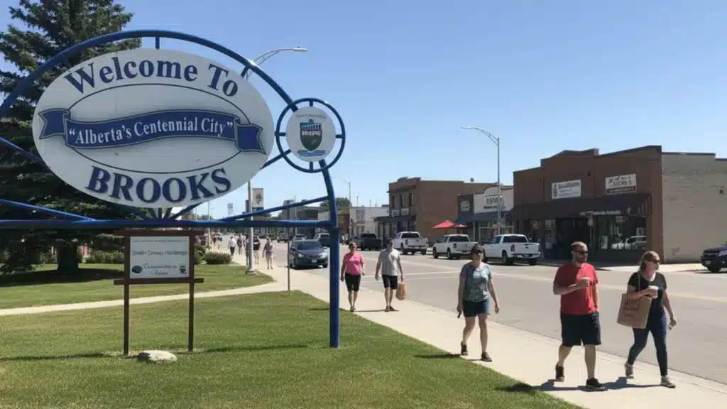People walk along a sunny street in Brooks Alberta, known for its business immigration and investment opportunities. A large sign reads “Welcome to BROOKS, Alberta’s Centennial City” as storefronts and cars line the road under a clear blue sky.