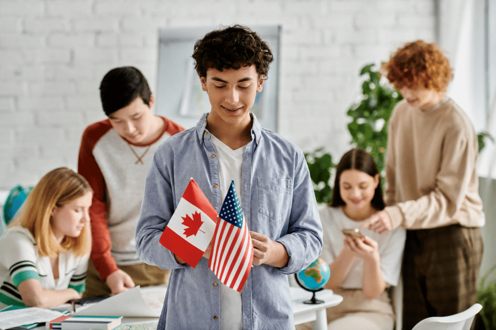 Young founder holding Canadian and American flags in a classroom as students collaborate behind him, weighing Canada vs US startup immigration.