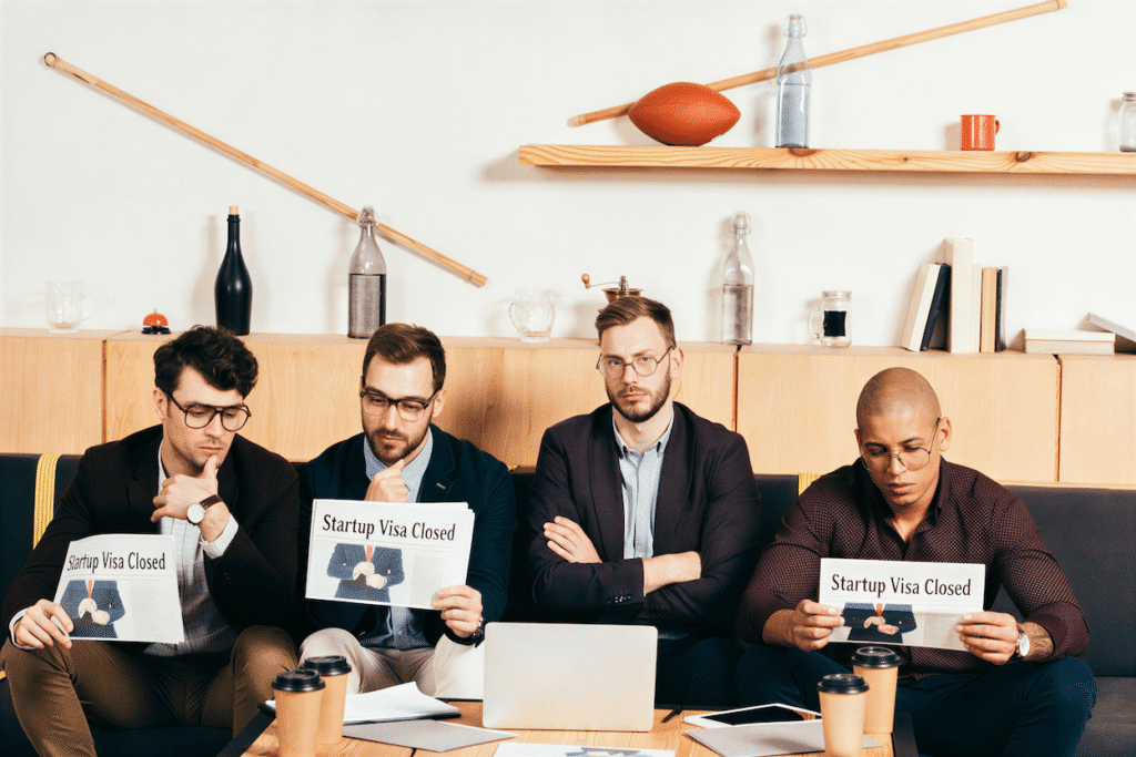Four men sit on a couch in an office, looking serious. Two are holding signs that say Startup Visa Closed, highlighting the ongoing issues with the Canada Start-Up Visa program. Coffee cups, papers, and a laptop sit on the table in front of them as they consider their options amid the growing Backlog 2026. Shelves with decor are in the background.